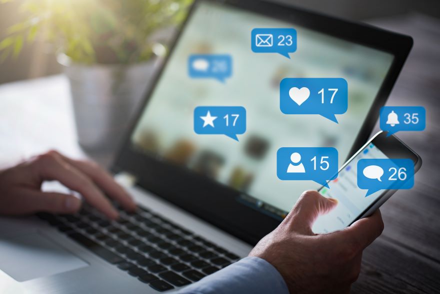 Hands typing on a laptop and holding a phone with social media interactions on screen