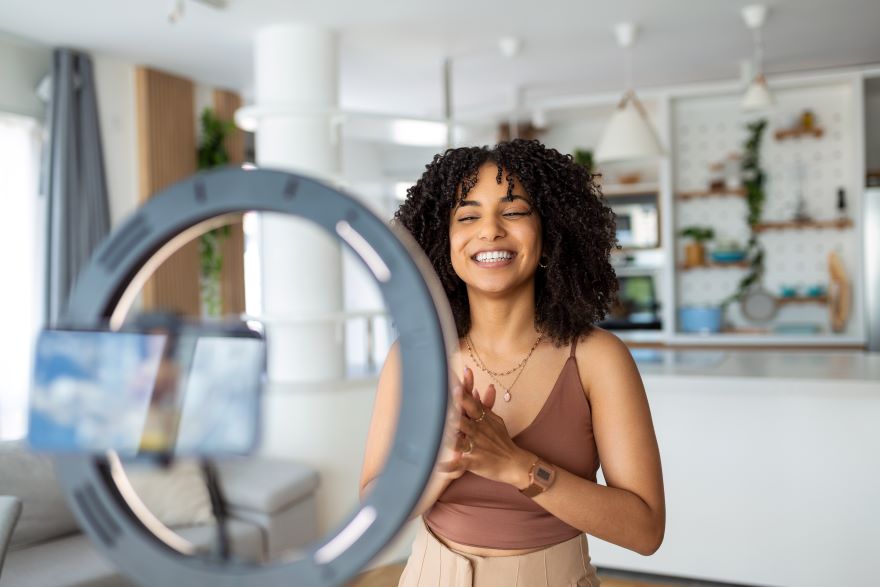 A woman with curly hair stands in front of a ring light, preparing to record a video in a modern, bright kitchen setting.