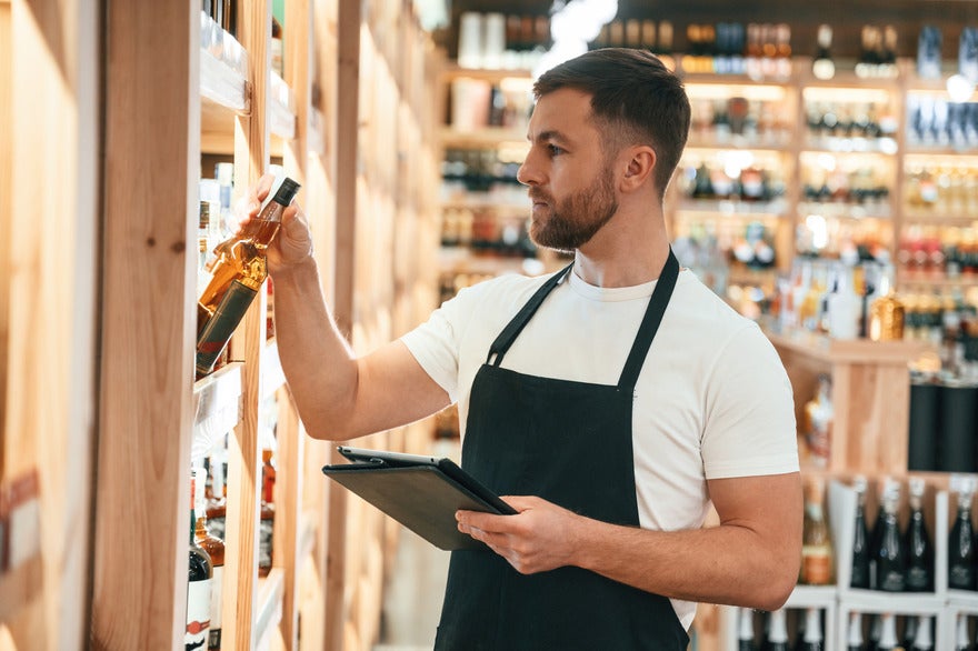 Male employee in an apron placing a bottle of alcohol on to a shelf
