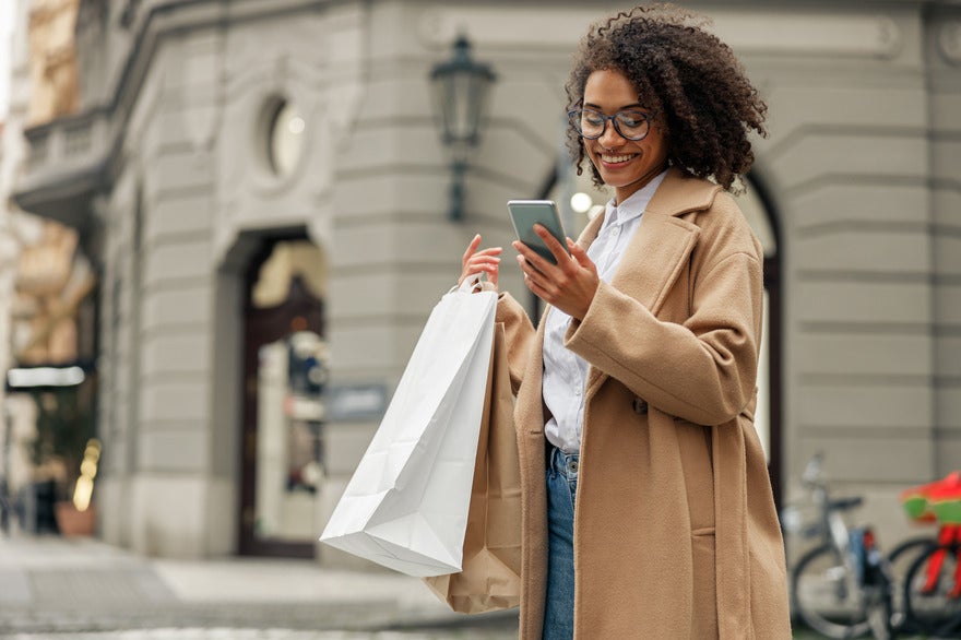 Woman in a coat standing outside with a couple of shopping bags while looking at her phone