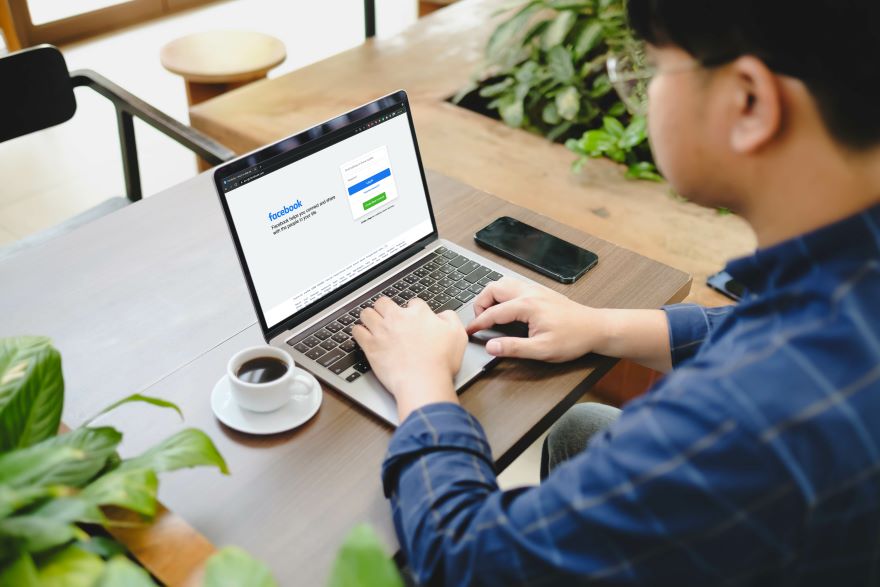 A person sits at a wooden table, using a laptop with the Facebook login page open, beside a cup of coffee and a smartphone.
