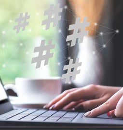 A person types on a laptop in a café, with floating hashtags above, symbolizing social media engagement. A cup is nearby.