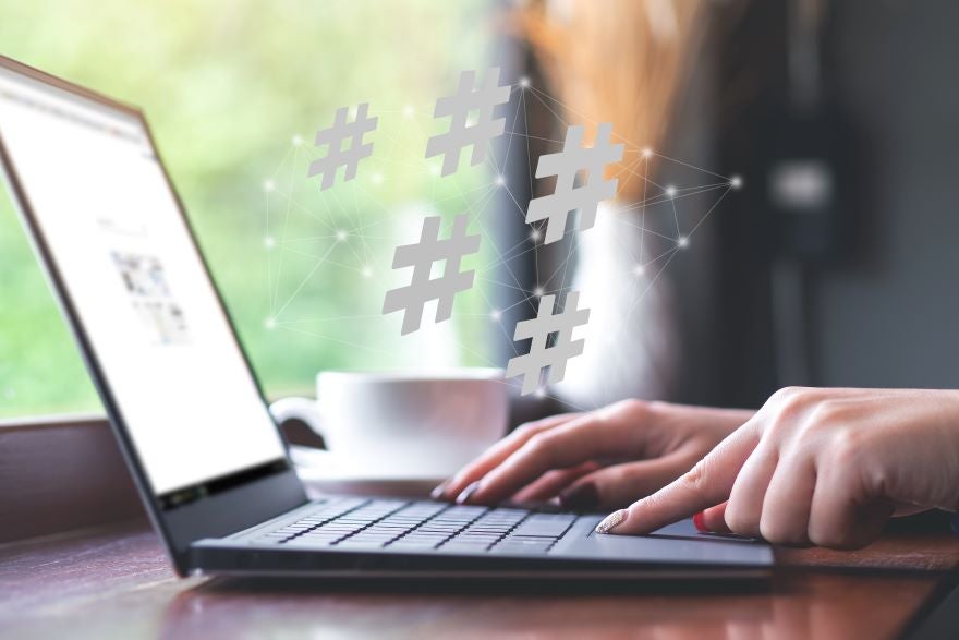 A person types on a laptop in a café, with floating hashtags above, symbolizing social media engagement. A cup is nearby.