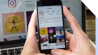 Hands hold a mobile phone showing Instagram's profile on Instagram, with a desktop behind also showing the same image