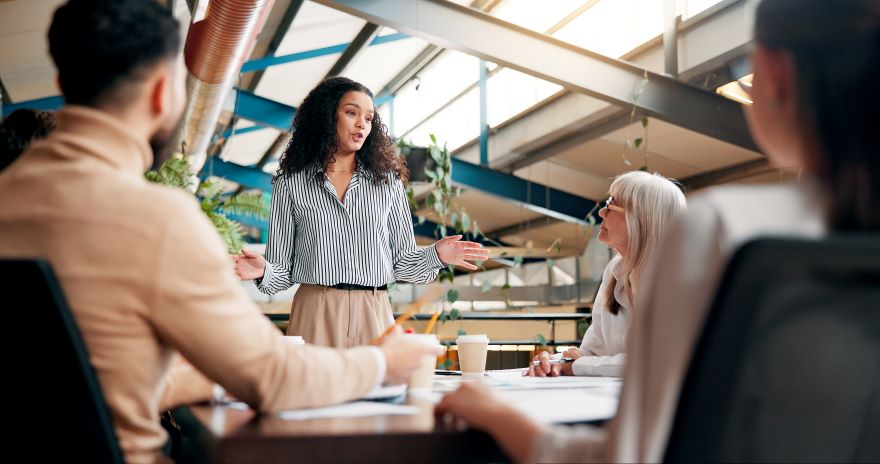 A woman in a striped shirt presents expressively in a bright meeting room, surrounded by attentive colleagues engaged in discussion.