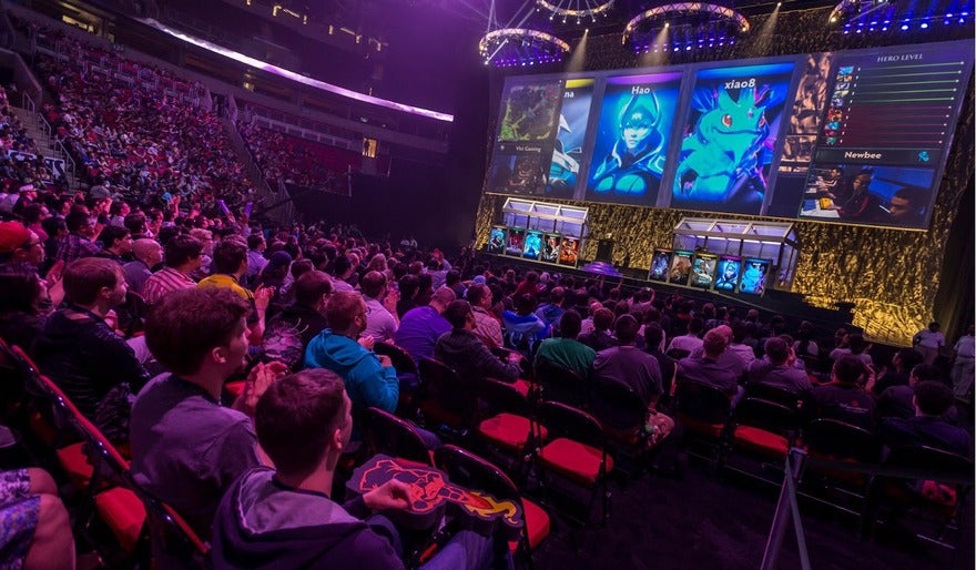 Audience watching an esports tournament with large screens displaying video game characters and player profiles.