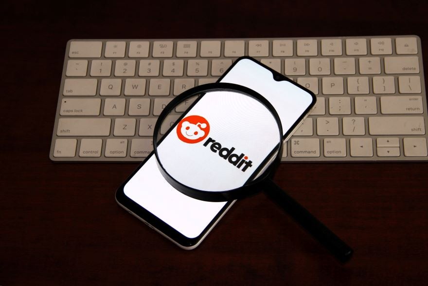 A smartphone displaying the Reddit logo, viewed through a magnifying glass, resting on a keyboard.
