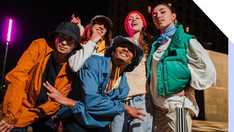 A group of five people in colorful streetwear poses playfully at night, with a vibrant pink light illuminating the background.