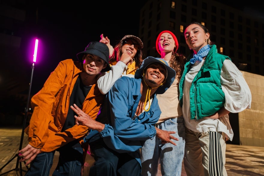 A group of five people in colorful streetwear poses playfully at night, with a vibrant pink light illuminating the background.