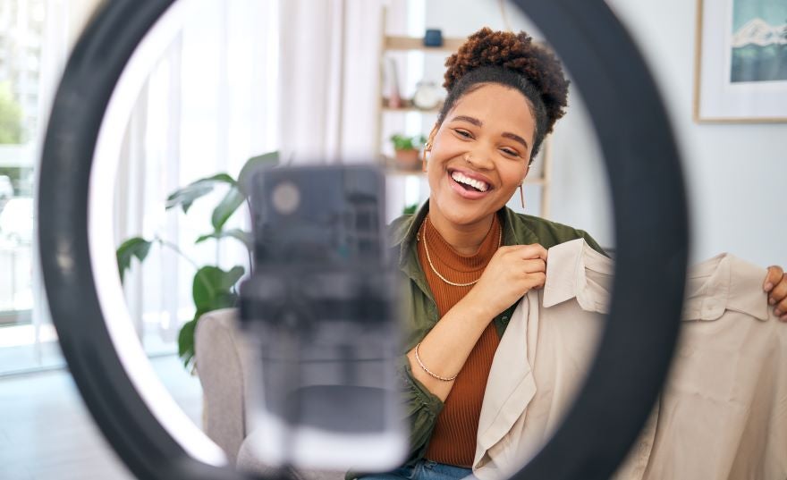 A woman smiling at a smartphone on a tripod holding up a shirt