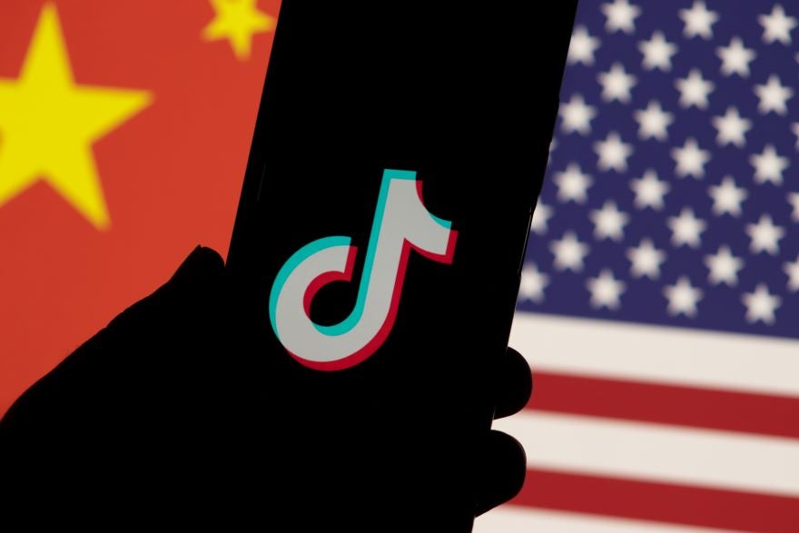 A hand holding a smartphone displaying the TikTok logo, with a background featuring the flags of China and the USA.