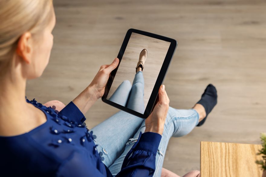 A person holds a tablet displaying a close-up of a shoe worn with jeans, sitting in a cozy, modern setting.